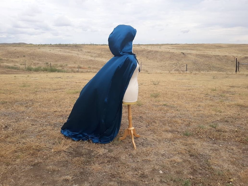 May include: A teal blue hooded cape with a satin finish. The cape is draped over a dress form and is displayed against a backdrop of a grassy field.