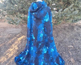 May include: A hooded cloak featuring a vibrant galaxy print with deep black, bright blue, and white star patterns. The garment has a full-length design that drapes downward from the shoulders, complete with an attached hood at the top.