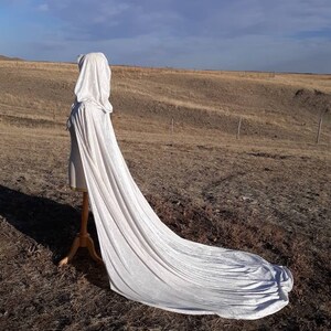 May include: A long white hooded cape with a flowing train, displayed on a mannequin against a backdrop of a brown field and a blue sky.