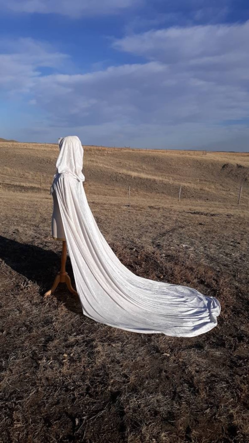 May include: A long white hooded cape with a flowing train, displayed on a mannequin against a backdrop of a brown field and a blue sky.