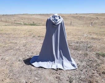 May include: A floor-length, solid light gray hooded cloak made of fabric. The garment features a structured hood at the top and drapes down to the ground in a wide, full-length silhouette, covering the entire back area of the display form.