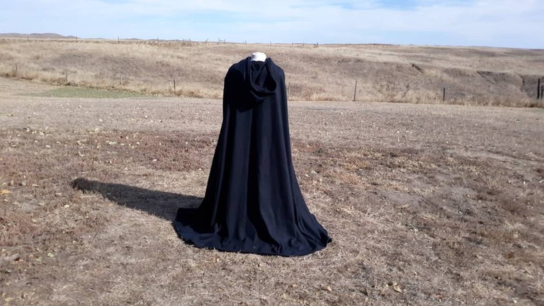 K&ouml;nnte beinhalten: Ein schwarzer Kapuzenumhang mit langem, flie&szlig;endem Design, fotografiert im Freien auf einem Feld.