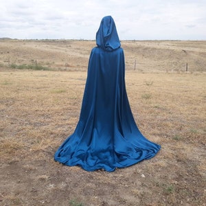 May include: A long, flowing blue hooded cape made of satin fabric. The cape is draped over a person standing in a field.