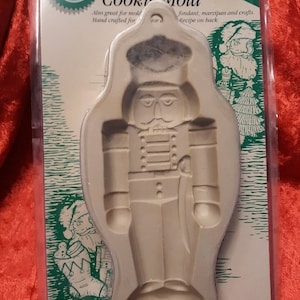 May include: A white plastic cookie mold shaped like a nutcracker. The mold is packaged on a white and green card with the text "Nutcracker Cookie Mold" and "Ovenproof!" The mold is also great for fondant, marzipan, and crafts.