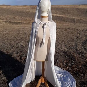 May include: A white velvet hooded cape with a tie closure. The cape is displayed on a mannequin against a brown grassy background.