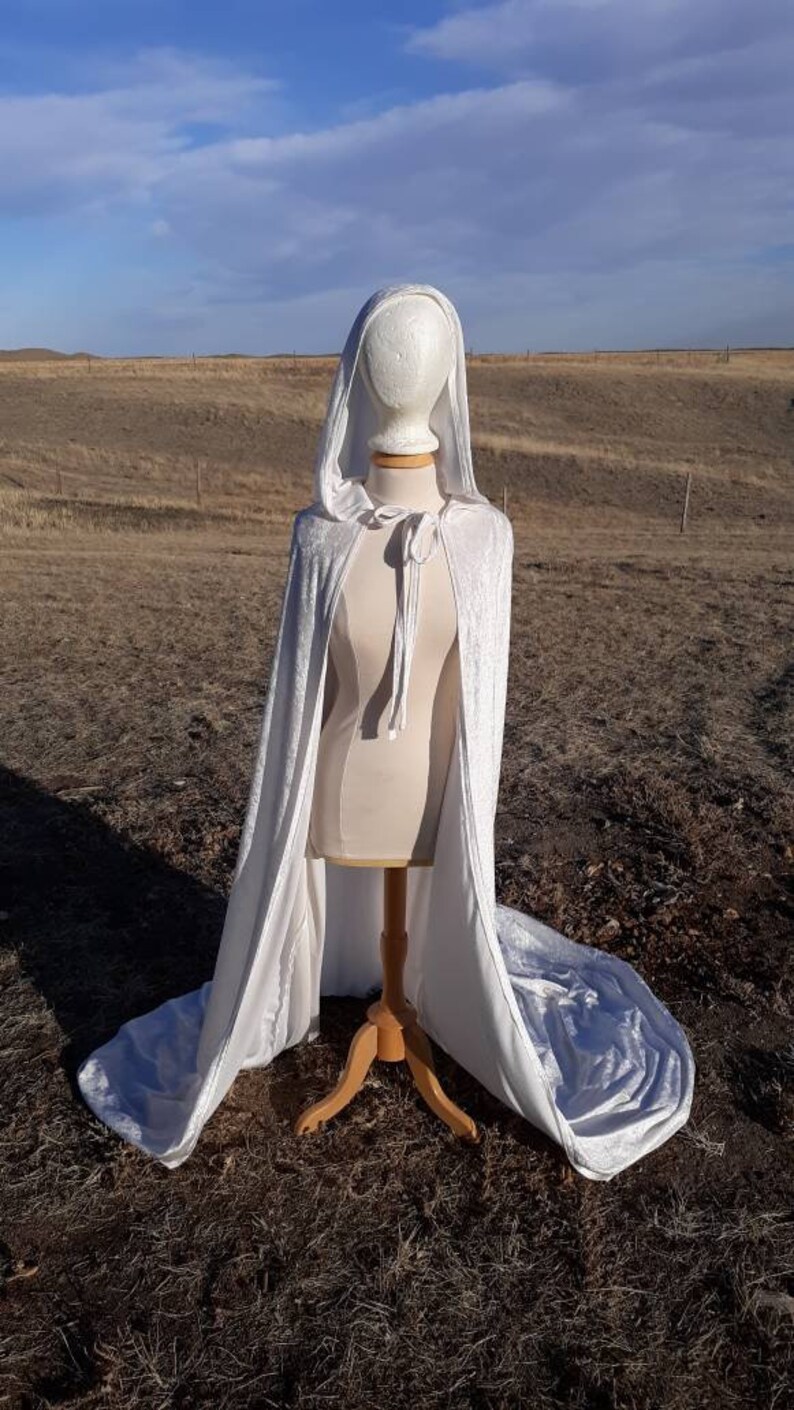May include: A white velvet hooded cape with a tie closure. The cape is displayed on a mannequin against a brown grassy background.