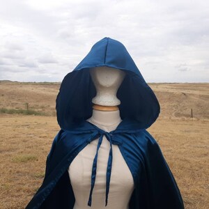 May include: A teal blue satin hooded cape with a tie closure. The cape is draped over a mannequin against a backdrop of a grassy field.