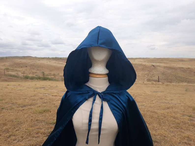 May include: A teal blue satin hooded cape with a tie closure. The cape is draped over a mannequin against a backdrop of a grassy field.