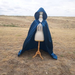 May include: A teal blue hooded cape with a satin finish. The cape is draped over a mannequin and is displayed against a backdrop of a grassy field.
