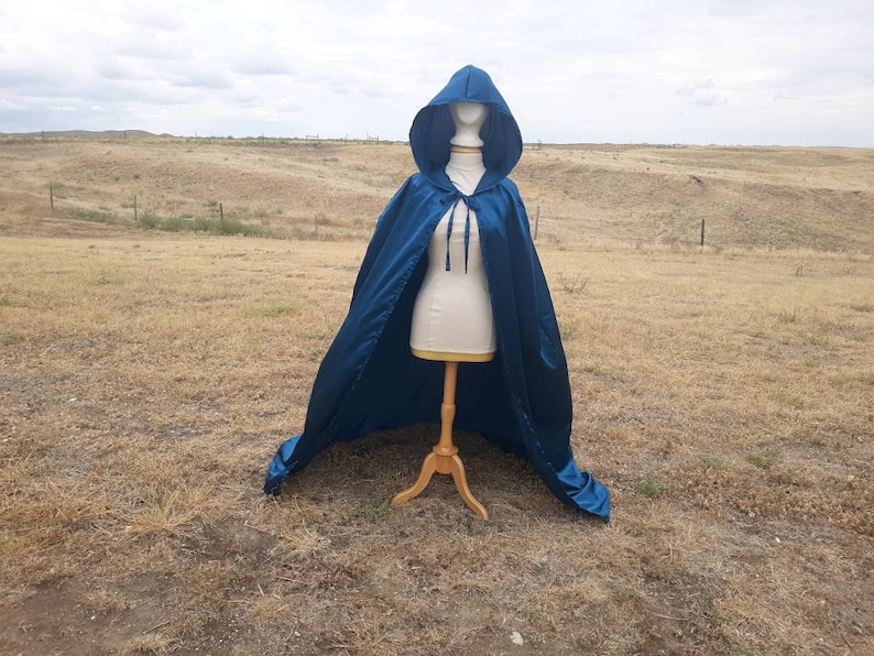 May include: A teal blue hooded cape with a satin finish. The cape is draped over a mannequin and is displayed against a backdrop of a grassy field.