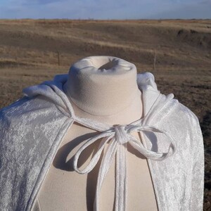May include: A white velvet hooded cape with a tie closure. The cape is displayed on a white mannequin against a backdrop of a brown field.