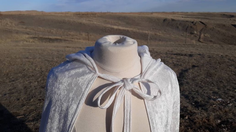 May include: A white velvet hooded cape with a tie closure. The cape is displayed on a white mannequin against a backdrop of a brown field.