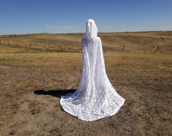 May include: A white lace hooded cape with a long train, photographed outdoors in a field.