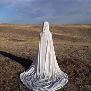 May include: A white hooded cape with a long train, made of velvet fabric, is displayed against a backdrop of a brown field and a blue sky with white clouds.