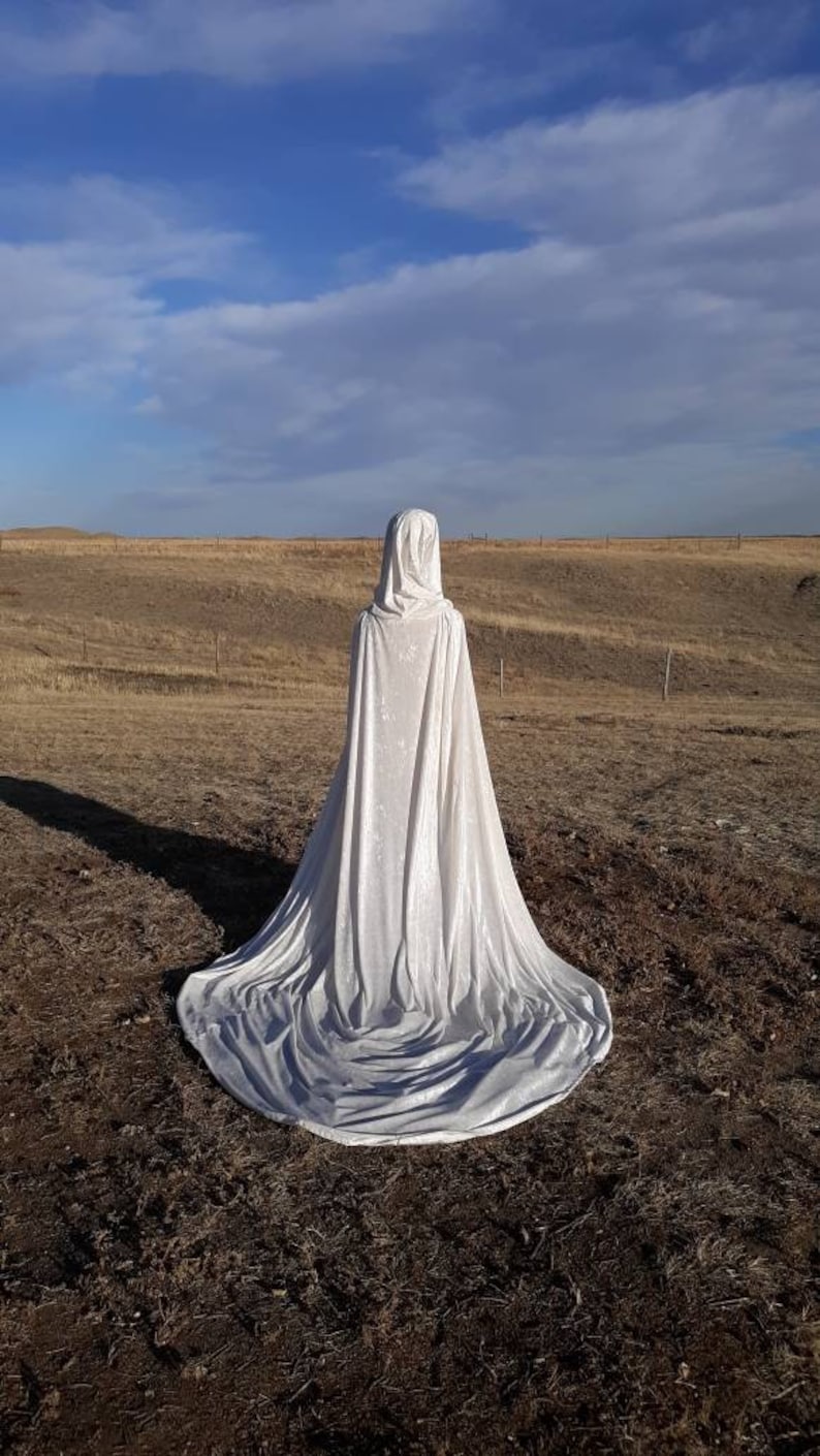 May include: A white hooded cape with a long train, made of velvet fabric, is displayed against a backdrop of a brown field and a blue sky with white clouds.