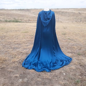 May include: A long, flowing blue hooded cape made of satin fabric. The cape is draped over a mannequin and is displayed against a backdrop of a grassy field.