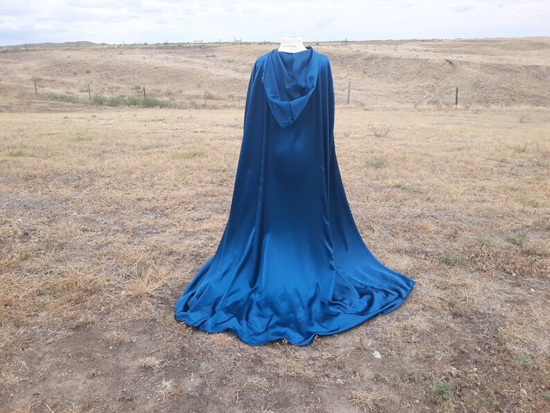 May include: A long, flowing blue hooded cape made of satin fabric. The cape is draped over a mannequin and is displayed against a backdrop of a grassy field.