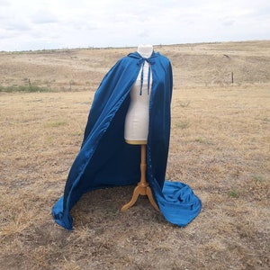 May include: A long, flowing teal blue cape with a hood and a tie closure. The cape is displayed on a mannequin in a field.