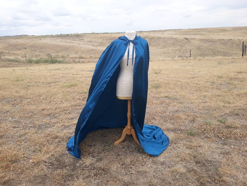 May include: A long, flowing teal blue cape with a hood and a tie closure. The cape is displayed on a mannequin in a field.
