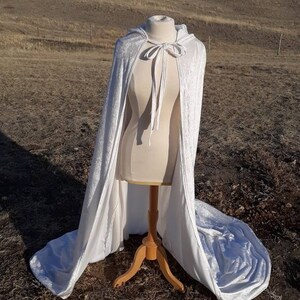 May include: A white velvet hooded cape with a tie closure. The cape is displayed on a wooden mannequin against a backdrop of a grassy field.