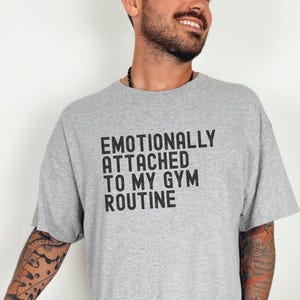 Könnte beinhalten: Sportgraues T-Shirt mit dem schwarzen Text "EMOTIONALLY ATTACHED TO MY GYM ROUTINE". Das Shirt ist von der Marke Gildan und wird mit einem braunen Football kombiniert. Das Shirt ist ein Freizeitartikel.