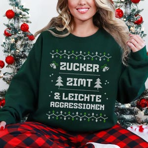 Könnte beinhalten: Ein dunkelgrünes Langarm-Sweatshirt mit einer weißen Grafik im Strick-Stil und dem Text ZUCKER ZIMT & LEICHTE AGGRESSIONEN, umgeben von dekorativen Mustern aus Weihnachtsbäumen, Glocken und Lichterketten in Weiß und Grün.