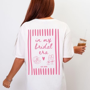 Könnte beinhalten: Weißes T-Shirt mit einem rosa Design auf der Rückseite. Das Design enthält den Text "in my bridal era", eine Ringbox, ein Herz und Sektgläser. Der Text "EST. 2026" ist ebenfalls vorhanden.