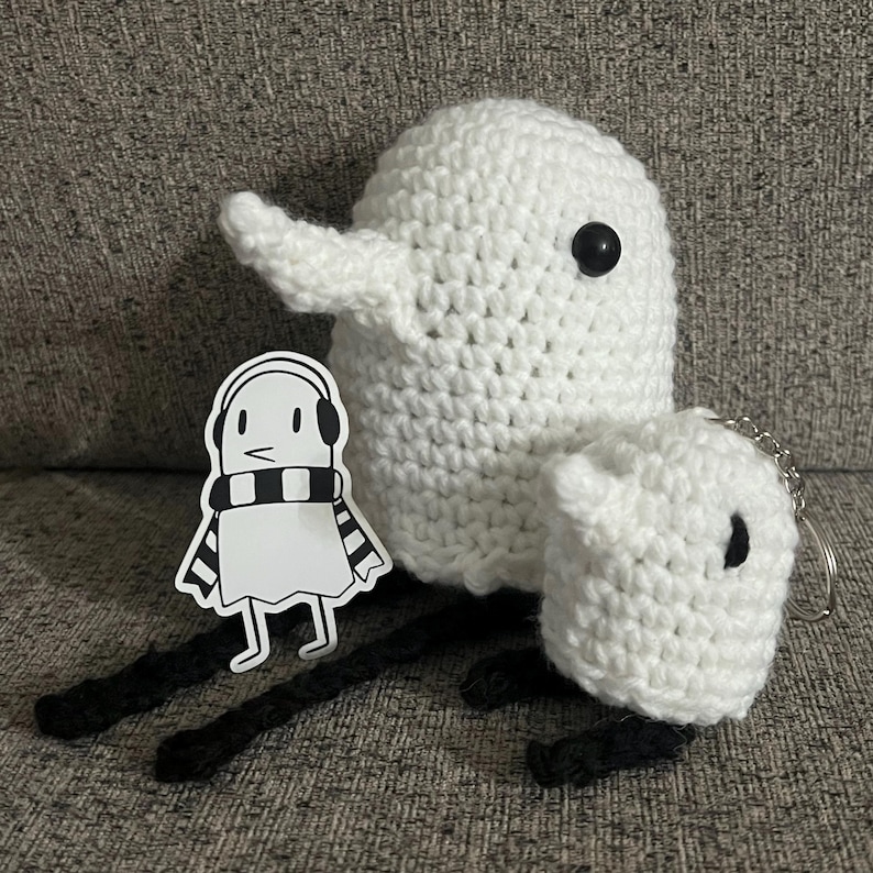 May include: Two white crocheted ghost plushies, one larger than the other, with black eyes. A black and white sticker of a ghost wearing a scarf is in the foreground.