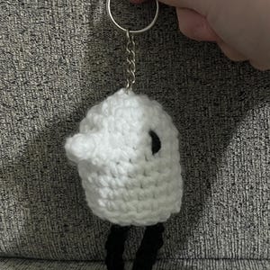 May include: White crocheted sheep keychain with black crocheted legs and a black eye. The keychain is attached to a silver key ring and chain.