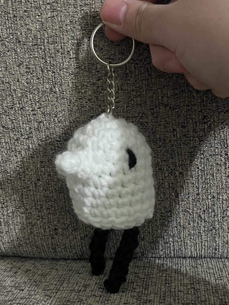 May include: White crocheted sheep keychain with black crocheted legs and a black eye. The keychain is attached to a silver key ring and chain.