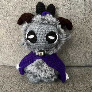 May include: A handmade crochet creature with gray fur, large black and white eyes, and brown horns. It wears a purple cape with white trim and a silver bell collar. The plush toy is designed to be a whimsical character.