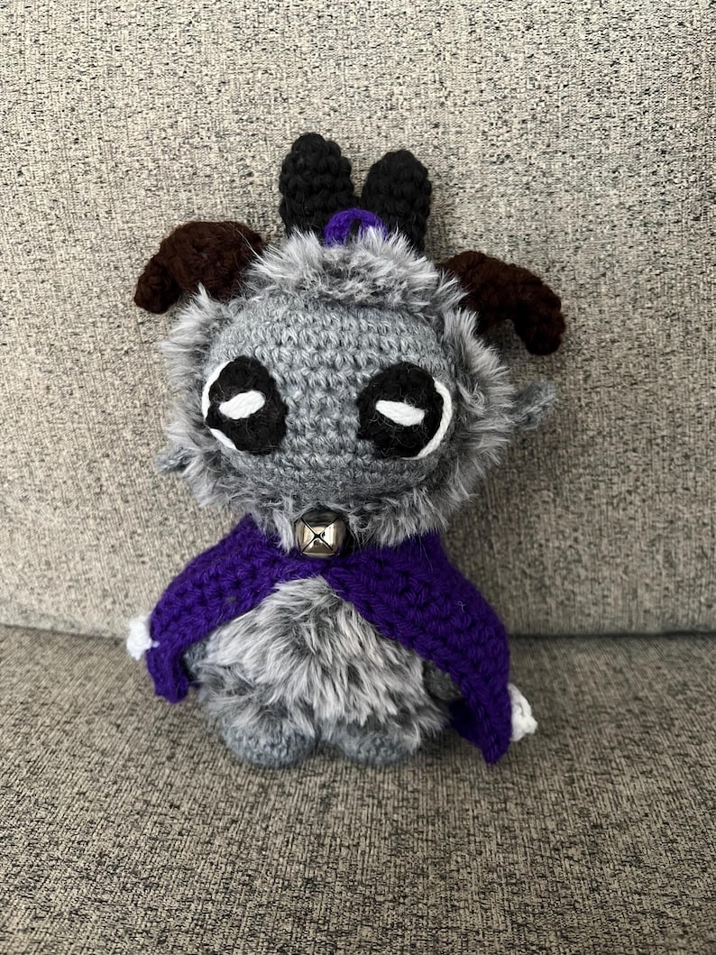 May include: A handmade crochet creature with gray fur, large black and white eyes, and brown horns. It wears a purple cape with white trim and a silver bell collar. The plush toy is designed to be a whimsical character.