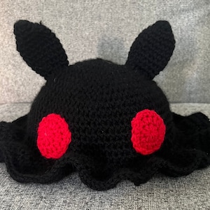 Puede incluir: Un gorro negro tejido a mano con dos orejas verticales y dos acentos circulares rojos. El gorro tiene un borde ondulado y es probablemente para un ni&ntilde;o o un adulto. El gorro est&aacute; hecho de hilo suave.