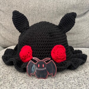 Puede incluir: Un sombrero negro de ganchillo con dos orejas peque&ntilde;as y dos c&iacute;rculos rojos. Una pegatina transparente de un murci&eacute;lago de dibujos animados con ojos rojos est&aacute; delante del sombrero. El sombrero tiene un borde con volantes.