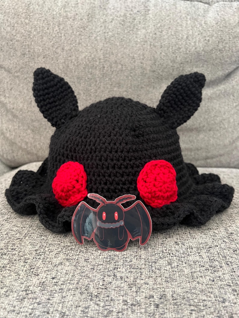 Puede incluir: Un sombrero negro de ganchillo con dos orejas peque&ntilde;as y dos c&iacute;rculos rojos. Una pegatina transparente de un murci&eacute;lago de dibujos animados con ojos rojos est&aacute; delante del sombrero. El sombrero tiene un borde con volantes.