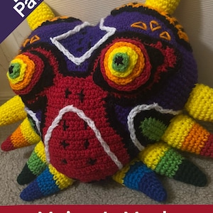 Könnte beinhalten: Eine gehäkelte Majora's Maske in Rot, Schwarz, Lila, Gelb, Weiß, Grün, Blau und Orange. Die Maske hat ein detailliertes Design mit mehreren Farben und Texturen. Das Bild enthält auch den Text "PDF Pattern" und "Majora's Mask Crochet Pattern".