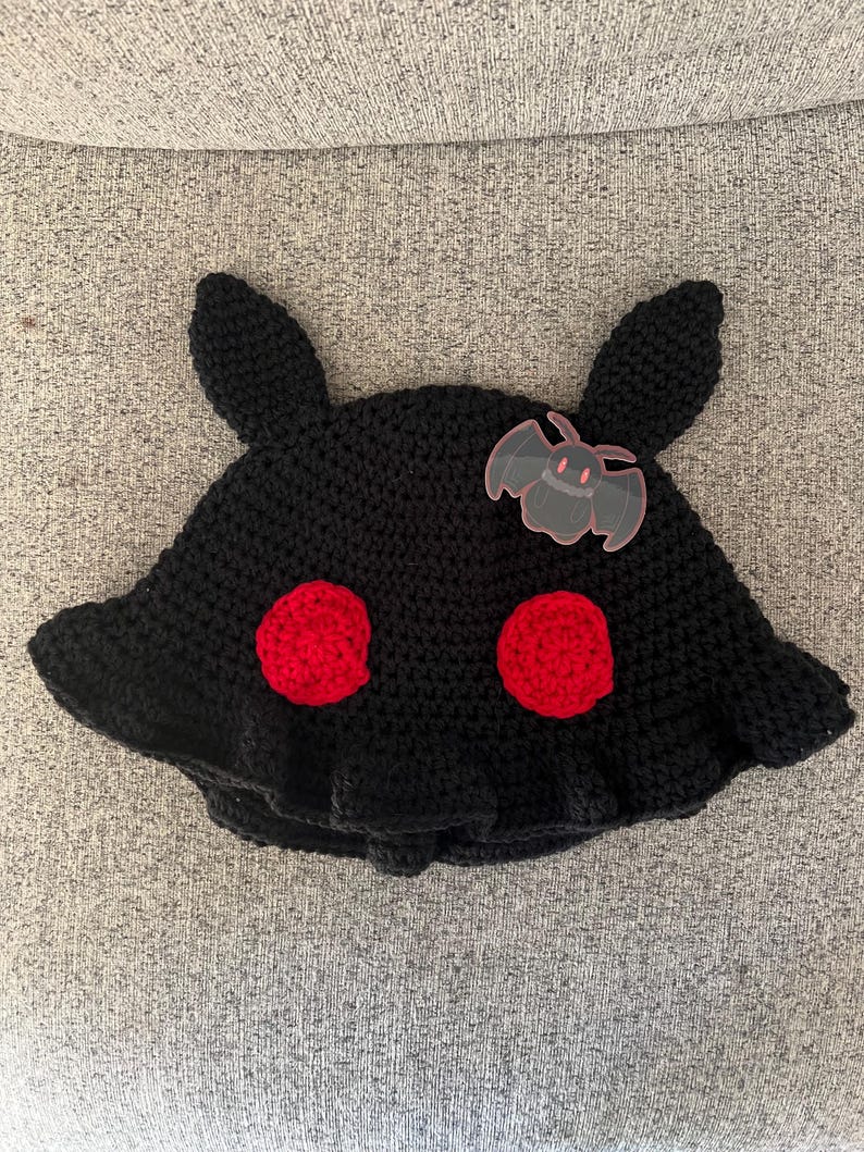 Puede incluir: Un sombrero negro de ganchillo con dos c&iacute;rculos rojos y dos orejas de conejo negras. Un peque&ntilde;o adorno transparente en forma de murci&eacute;lago est&aacute; adherido al sombrero. El sombrero est&aacute; sobre una superficie gris.