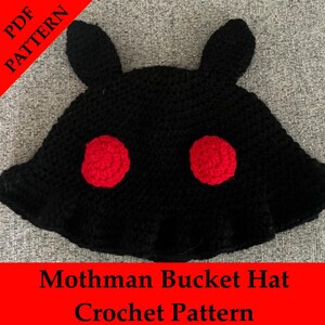 Może przedstawiać: Czarny szydełkowy kapelusz typu bucket z dwoma czerwonymi okręgami i dwoma kształtami przypominającymi uszy. Na kapeluszu widnieje napis "Mothman Bucket Hat Crochet Pattern" na czerwonym banerze. Ręcznie robiony.