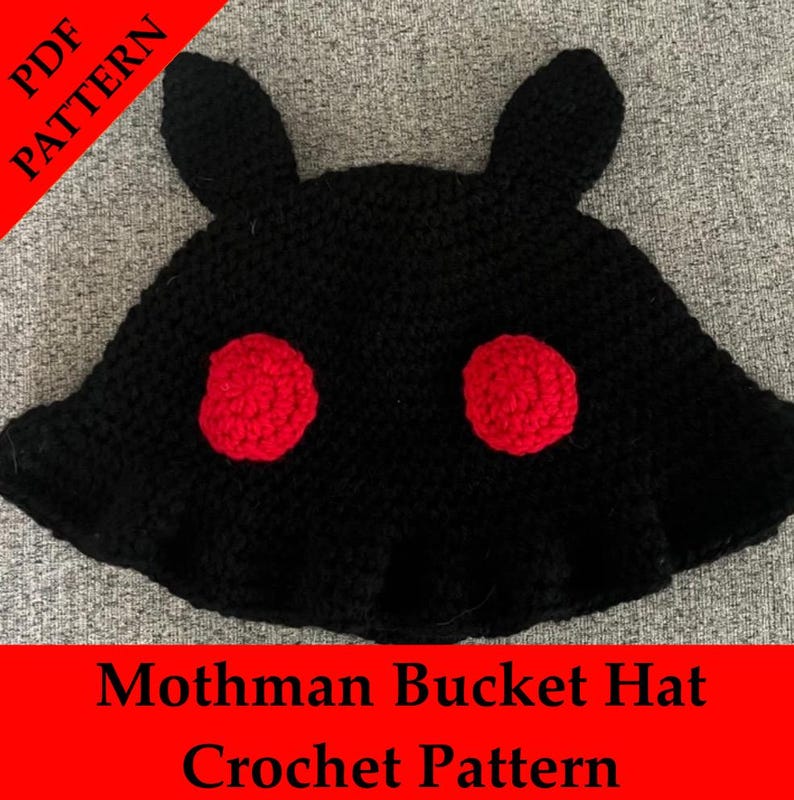 Może przedstawiać: Czarny szydełkowy kapelusz typu bucket z dwoma czerwonymi okręgami i dwoma kształtami przypominającymi uszy. Na kapeluszu widnieje napis "Mothman Bucket Hat Crochet Pattern" na czerwonym banerze. Ręcznie robiony.
