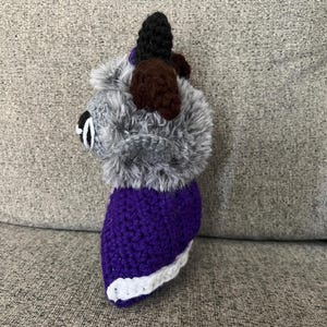 May include: A handmade crochet dog toy with a gray fluffy head, brown ears, and a black top. The body is purple with a white trim. The toy is sitting on a gray surface.