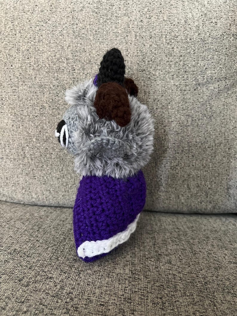 May include: A handmade crochet dog toy with a gray fluffy head, brown ears, and a black top. The body is purple with a white trim. The toy is sitting on a gray surface.