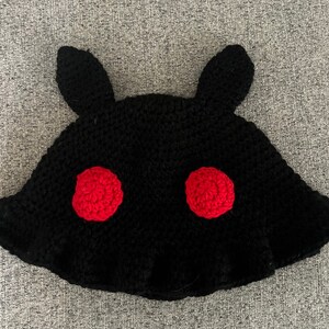 Puede incluir: Un sombrero negro de ganchillo con dos protuberancias verticales en forma de orejas y dos grandes acentos circulares rojos. El sombrero est&aacute; hecho a mano y parece ser una pieza de disfraz o un art&iacute;culo de novedad. El sombrero est&aacute; sobre una superficie gris.