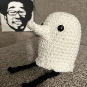 May include: White crocheted ghost with black yarn legs and black button eyes. The ghost is sitting on a gray surface.