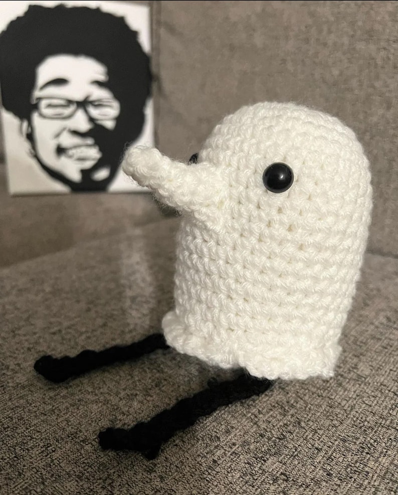 May include: White crocheted ghost with black yarn legs and black button eyes. The ghost is sitting on a gray surface.