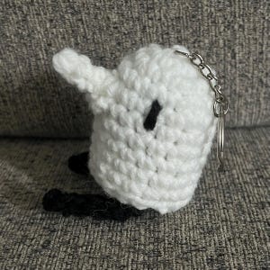 May include: White crocheted keychain with black details shaped like a ghost with a small black eye. The keychain has a silver metal ring.