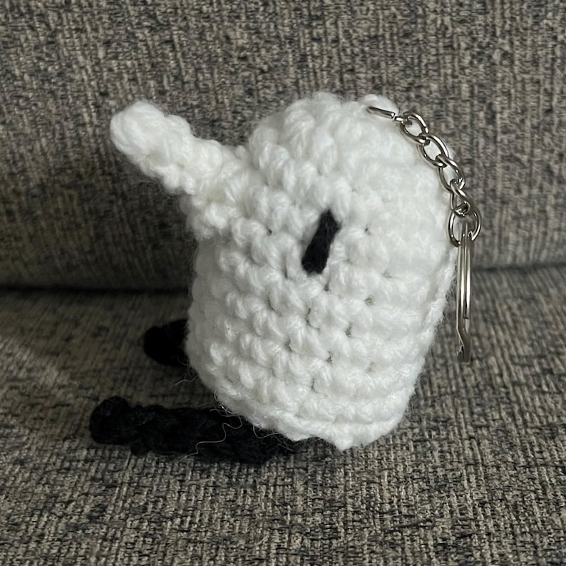 May include: White crocheted keychain with black details shaped like a ghost with a small black eye. The keychain has a silver metal ring.