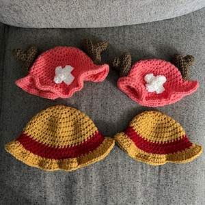 May include: Two pink crocheted hats featuring brown antlers and a white cross emblem, alongside two yellow crocheted bucket hats with a red band around the base. These four handmade items are crafted with a textured yarn stitch pattern throughout.
