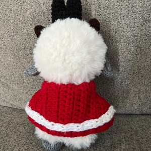 May include: A handmade plush toy with a fluffy white body, black antlers, and gray ears and feet. It wears a red crocheted cape with white trim. The toy is designed to resemble a reindeer.