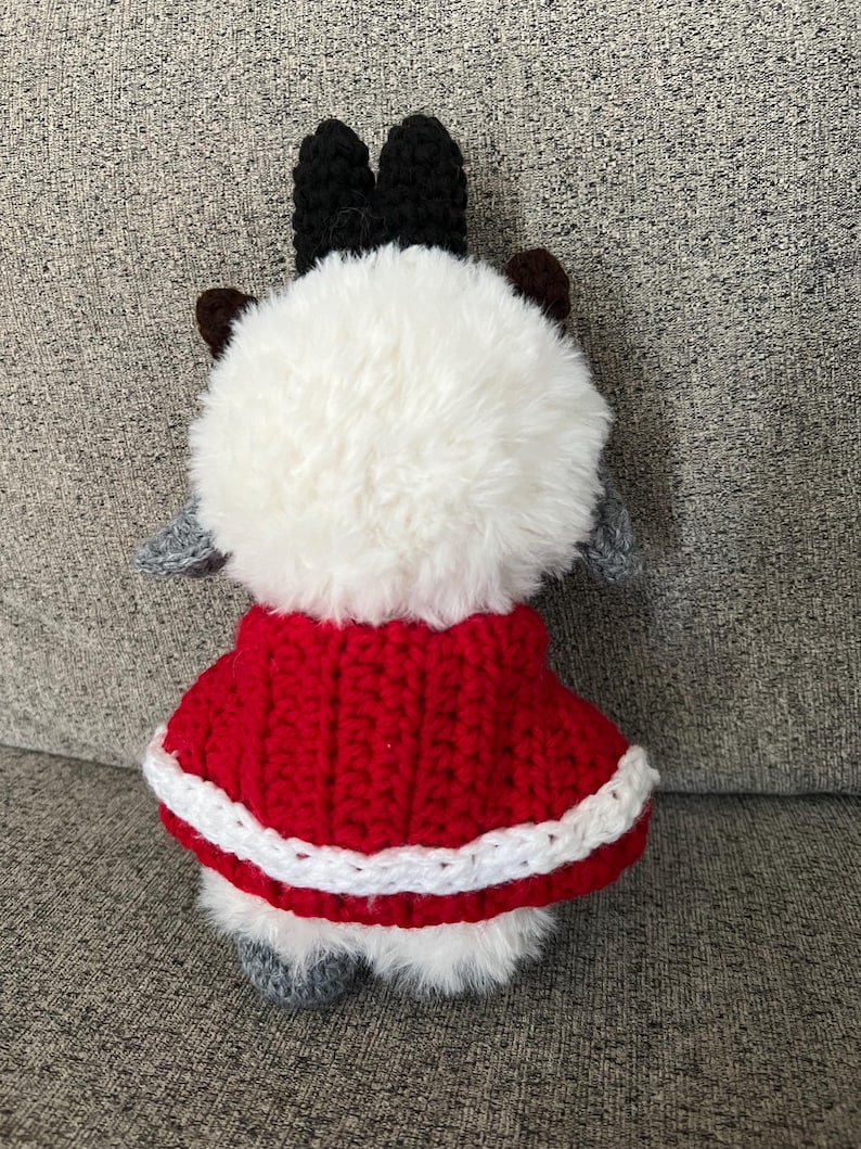 May include: A handmade plush toy with a fluffy white body, black antlers, and gray ears and feet. It wears a red crocheted cape with white trim. The toy is designed to resemble a reindeer.