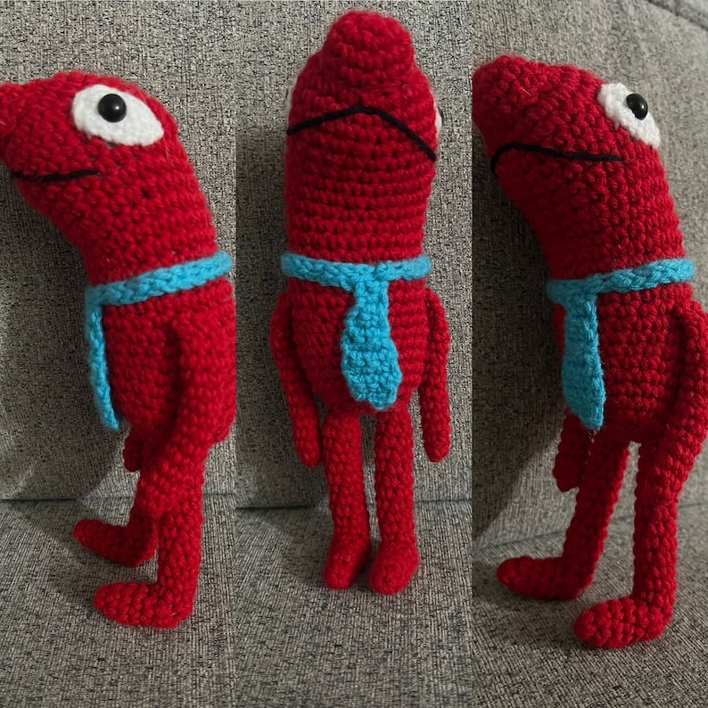 Op de afbeelding: Drie gehaakte rode figuren met blauwe stropdassen. De figuren hebben witte ogen en staan op een grijs oppervlak.